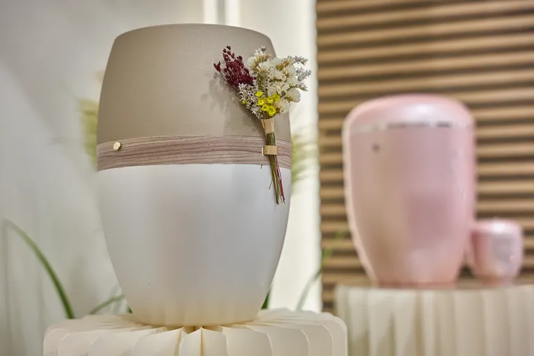 Urne in Taupe und Weiß mit Trockenblumen-Dekor, im Hintergrund eine rosafarbene Urne